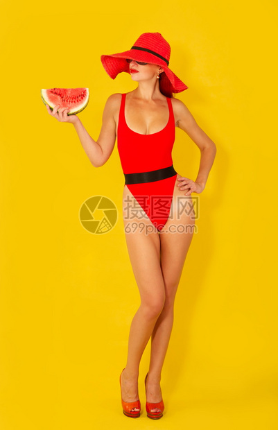 年轻女孩穿着明亮的红色泳衣和宽的红帽子在黄色背景上吃西瓜图片