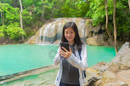 在泰公园旅行和度假期间快乐的亚洲女使用移动智能手机在热带森林的Erawan瀑布上聊天或致电社交媒体朋友图片