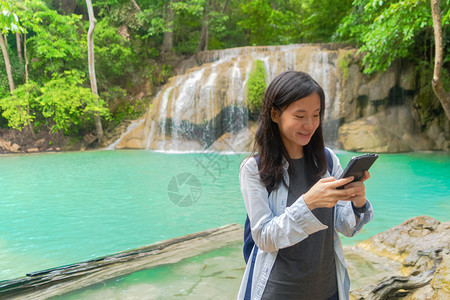 在泰公园旅行和度假期间快乐的亚洲女使用移动智能手机在热带森林的Erawan瀑布上聊天或致电社交媒体朋友图片