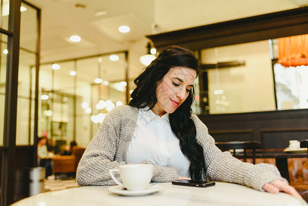 漂亮的黑发女人检查她的智能手机边吃早餐在旧馆喝咖啡图片