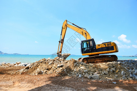 在海滨洋和蓝天空背景的建筑工地背鞋装载器上工作的挖土机掘石图片