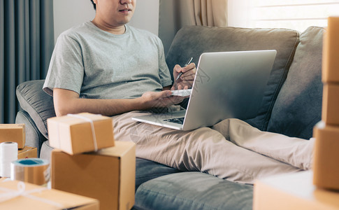 坐在沙发上的年轻亚洲男子在看电脑笔记本和纸上写单中小企业概念图片