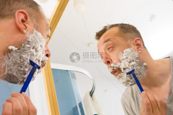 脸上有很多剃须泡沫的男人站在浴室里看着镜子刮胡图片