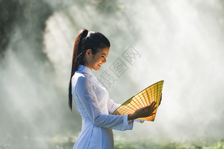 亚洲妇女维特南或中华风格的服装美丽年轻女孩外衣穿着传统服装的肖像在自然背景上手握着帽子编织叶图片