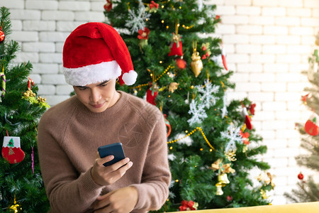 穿着棕色毛衣的年轻亚洲成男孩使用手机为社交媒体提供手机等待女孩朋友在带有圣诞节装饰的客厅里庆祝圣诞节背景图片