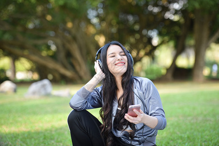 亚洲女用手机在户外听音乐快的年轻女孩微笑放松运动在绿色花园公用智能手机耳收听音乐图片