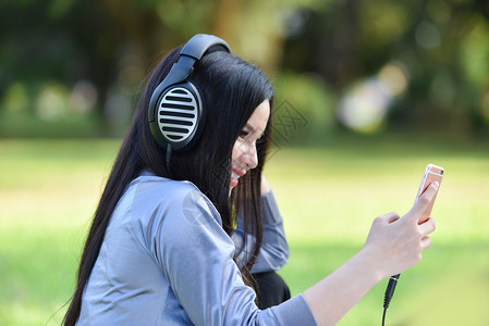 亚洲女用手机在户外听音乐快的年轻女孩微笑放松运动在绿色花园公用智能手机耳收听音乐图片