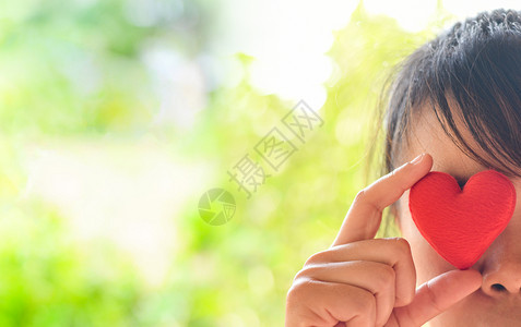 亚裔女快乐的年轻孩怀着红心闭眼睛看大自然的绿色bokeh模糊的背古在爱的情人节和慈善概念中图片