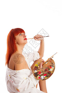 一个穿着涂漆烟雾衬衫的年轻红头发女孩坐在一个有纸垫的挂着一个带调色板和刷子的空帆布挂板前背景图片