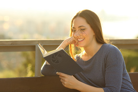 快乐的女人在读一本有趣的书日落时坐在户外的长椅上图片