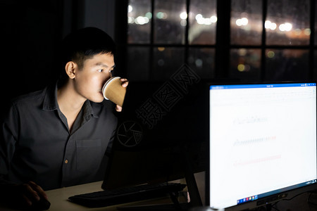 年轻成亚洲商人在办公室深夜使用台式电脑工作时喝热咖啡图片