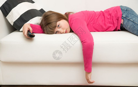 一个无聊的小女孩躺在沙发上看电视在科罗纳检疫期间背景图片