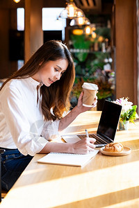 身着智能散装服的年轻亚洲女在客厅工作使用笔记本电脑在家工作喝热咖啡和羊角面包图片