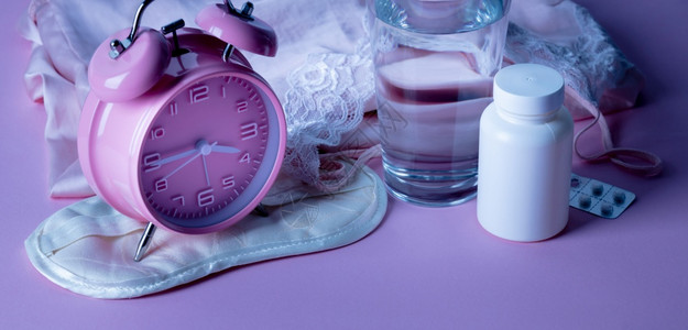 玻璃奶瓶粉红色背景的闹钟眼罩药丸和水杯失眠或无睡夜晚的抑郁治疗闹钟药丸和粉红色背景的水杯背景