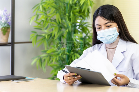 Asian女医生的肖像是戴保护面罩的亚洲女医生坐在院诊所的办公室里在检查耐心之前先阅读文件然后检查耐心图片