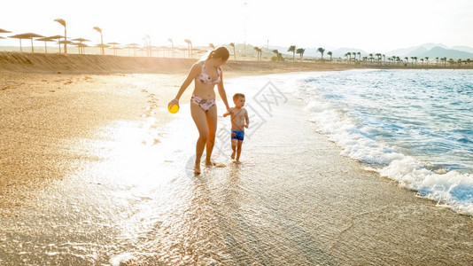 快乐的年轻母亲用手抱着她小孩的美丽形象在寒冷的海滨上行走与惊人的日落天空相对的温暖平静波浪相对美丽的年轻母亲用手抱着她小孩的美丽图片