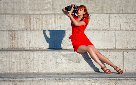 穿着亮红礼服的年轻美女摄影师专注于一个配有大型数码相机的模特图片