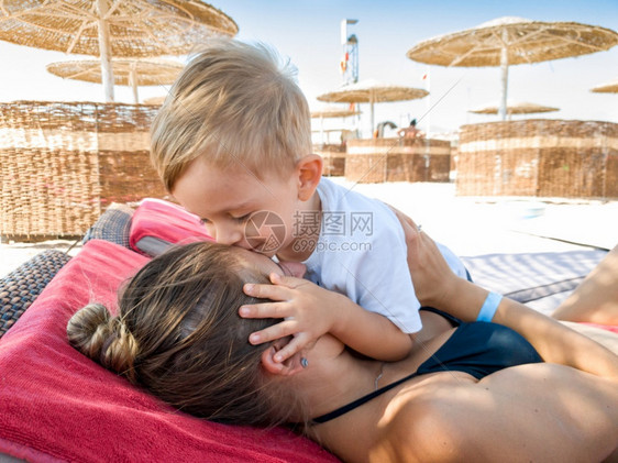 3岁可爱的男孩肖像亲吻他母在阳光保护伞下海边放松家庭在暑假期间沙滩上放松和愉快3岁可爱的幼儿男孩肖像在阳光保护伞下海边放松图片