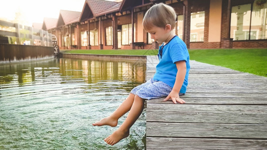 照片来自三岁可爱的男孩坐在河岸上老城的水渠上并用脚泼水图片