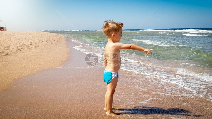 站在沙海滨用食指对准地平线的可爱幼儿男孩肖像图片