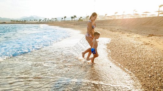 照片快乐的年轻母亲与可爱的3岁幼儿男孩手牵在海滨与水面美丽的日落天空对面行走图片快乐的年轻母亲与可爱的3岁幼儿男孩手牵在海边与美图片