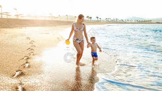 照片快乐的年轻母亲与可爱的3岁幼儿男孩手牵在海滨与水面美丽的日落天空对面行走图片快乐的年轻母亲与可爱的3岁幼儿男孩手牵在海边与美图片
