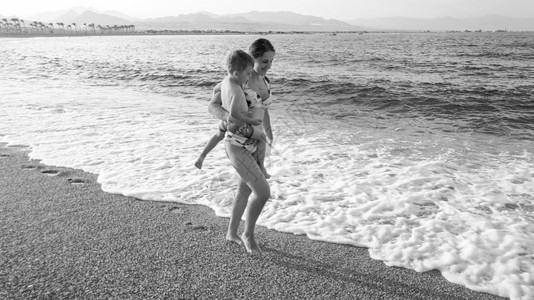 年轻女拥抱其小儿子在海边漫步宁静的温暖波浪中平静地走在海边的年轻女黑白照片子女和父母在海滩上放松年轻女拥抱其小儿子在海边平静地走图片