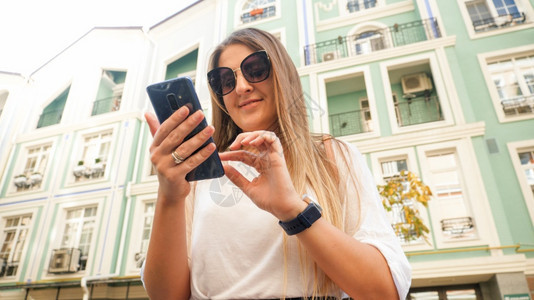 阳光眼镜中的年轻美女肖像在城市街道的智能手机上打字图片