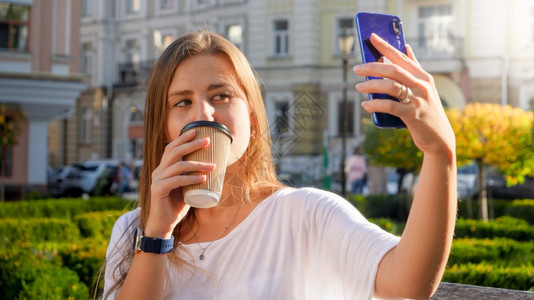 美丽的年轻女人肖像她用咖啡杯装扮同时在智能手机上自拍图片