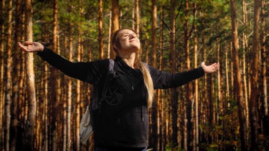 美丽的微笑女游客肖像背包伸展的手在松树林中仰望明亮的太阳自由概念和与然谐相处的概念背包伸展的手在松树林中仰望明亮的太阳自由以及与图片