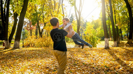 年轻父亲在秋天公园抱着和旋转自己的小儿子图片
