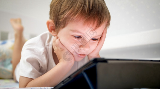 穿着睡衣躺在床上并使用数字平板电脑的微笑小男孩肖像图片