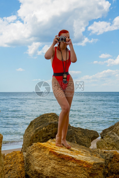一个穿着紧闭红色泳衣的年轻女孩带着一个反光相机和测量仪站在岩石海滩上拍照图片
