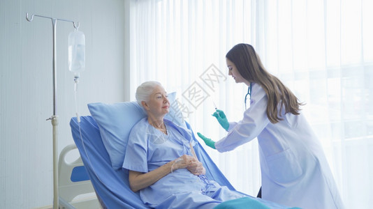 快乐的女医生为病老年女病人提供注射针头疫苗她们躺在医院病房的床上接受医疗和治图片