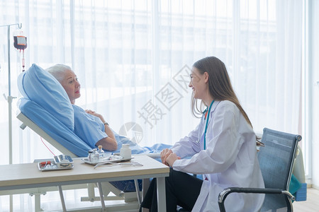 快乐的女医生讲话鼓励病的老年女病人躺在医院房的床上接受医疗和图片
