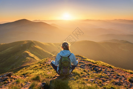 风景和人穿着背包坐在山峰上的年轻女子和美丽的山峰在夏季日落时雾中与运动女郎绿林山丘天空太阳光旅行和游瑜伽背景