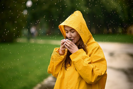 穿着雨衣的女子在天夏季公园喝热茶独自一人在走道上披着雨斗篷的女子在巷里的湿天气中穿雨衣的女子在公园喝热茶雨天穿衣的女子喝热茶图片