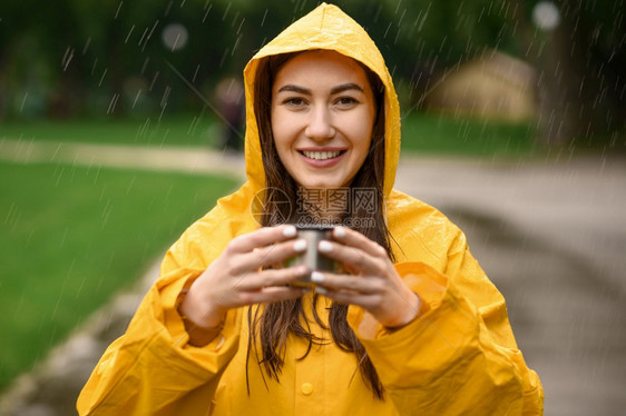 穿雨衣的妇女拿着热茶杯在雨天的夏日公园透过雨滴看一独自人在走道上披着雨斗篷的女在巷子里的湿天气穿雨衣的妇女拿着杯子透过滴水看一图片