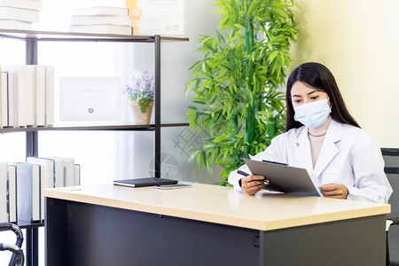 Asian女医生的肖像是戴保护面罩的亚洲女医生坐在院诊所的她办公室里在检查耐心之前先阅读文件然后检查耐心图片