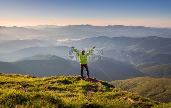 在山峰上站着运动男子手举起来对抗山谷秋天的日落雾中年轻男子岩石森林和蓝天图片