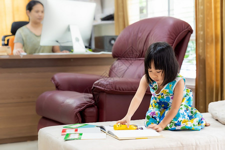 亚洲女童在家里客厅阅读互动式书作为家庭教育而她的母亲则在家里工作因为城市由于共食19大流行而被封锁背景图片