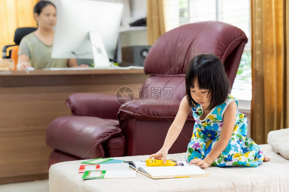 亚洲女童在家里客厅阅读互动式书作为家庭教育而她的母亲则在家里工作因为城市由于共食19大流行而被封锁图片