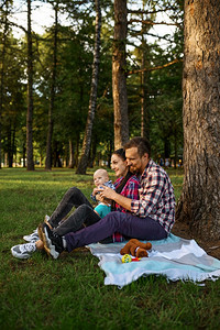父母亲和小婴儿坐在夏天公园的树下父母和男孩在草坪上与子一起在森林中野餐家庭幸福母亲父和婴儿坐在树下图片