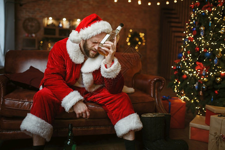 糟糕的圣诞老人和酒瓶坐在沙发上宿醉不健康的生活方式在节日服装新年和酗酒中留胡子的男坏圣诞老人和在沙发上装酒图片