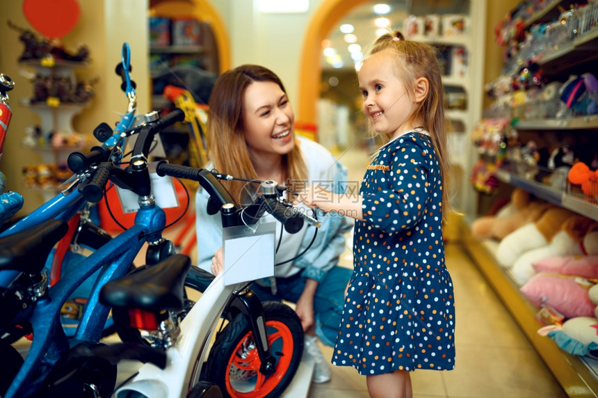 母亲和可爱的女儿在玩具店快乐的童年家庭在商店购买妈和小女孩选择自行车子和商店图片