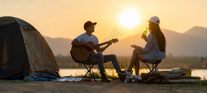 亚洲成年夫妇在日落时帐篷露营地边弹吉他和喝啤酒图片