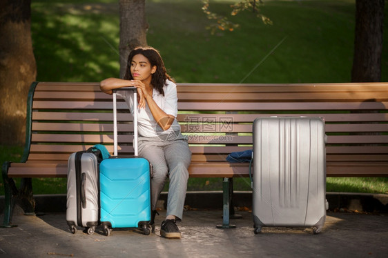 在夏季公园的长椅上坐着手提箱的年轻妇女在户外随身携带行李闲暇的女旅者在大自然中随身携带行李包的乘客在城市小巷上携带行李的女童在公图片