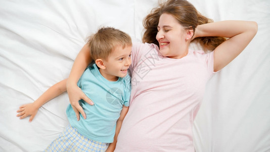 快乐的年轻母亲和穿着睡衣躺在床上微笑着的小孩儿子首观父母亲和家庭幸福感的首观快乐的年轻母亲和穿着睡衣躺在床上笑着的小孩儿子首观养图片