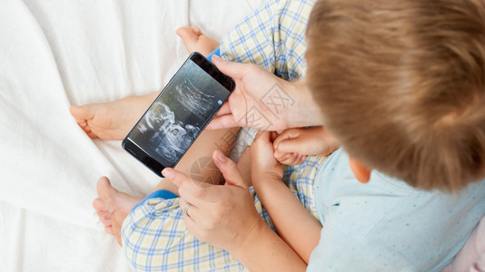 年轻女通过智能手机向小儿子展示婴超声波图像医疗保健和家庭幸福预期婴儿的概念医疗保健和家庭幸福预期婴儿概念图片