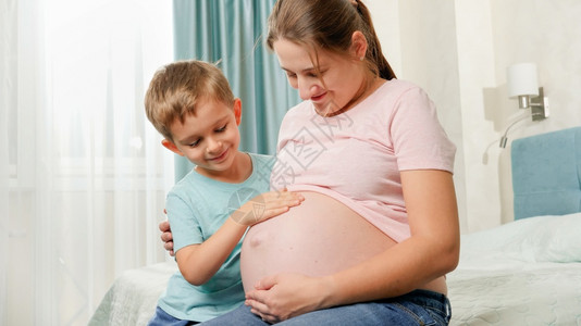 坐在卧室的怀孕母亲肚子上家庭期望婴儿的情绪积极可爱的男孩在卧室床上感受到怀孕母亲的大肚子图片